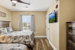 A bedroom with a bed, a window with curtains, a ceiling fan, a wall-mounted TV, and a wooden dresser.