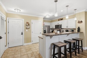 Una cocina con armarios blancos, un refrigerador de acero inoxidable, un microondas y una encimera de granito. Hay tres taburetes de bar y luces colgantes sobre la encimera.