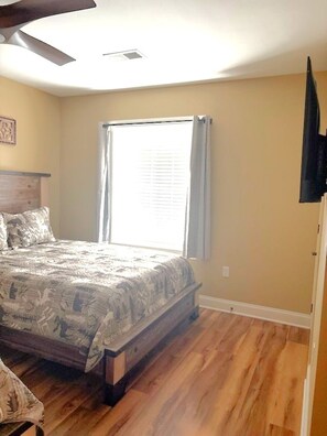 A bedroom with a bed, a ceiling fan, a window with curtains, and a wooden floor.