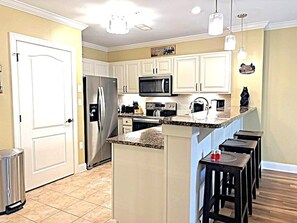 A kitchen with white cabinets, a stainless steel refrigerator, a microwave, and a granite countertop island with bar stools.