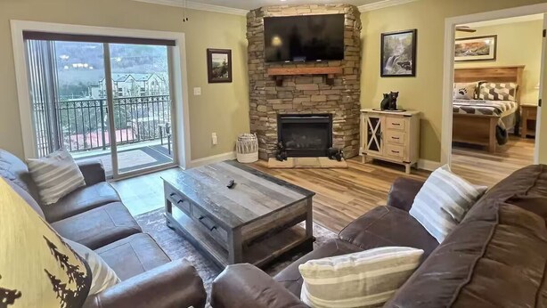 A living room with a stone fireplace, a flat-screen TV, a ceiling fan, and a large window overlooking a balcony.