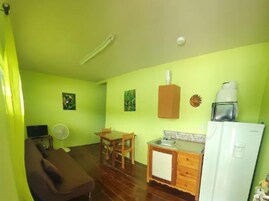 Una habitación pequeña e iluminada con una mesa de comedor de madera, un sofá, un refrigerador blanco y una pequeña zona de cocina con un lavamanos y una cafetera.