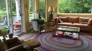 A cozy living room with a brown leather sofa, a grey armchair, and a coffee table with books and a lamp. Large windows offer a view of greenery outside. - Charming Home, Panoramic Mountain Views, Ideal For Family (Bolton-Ouest)