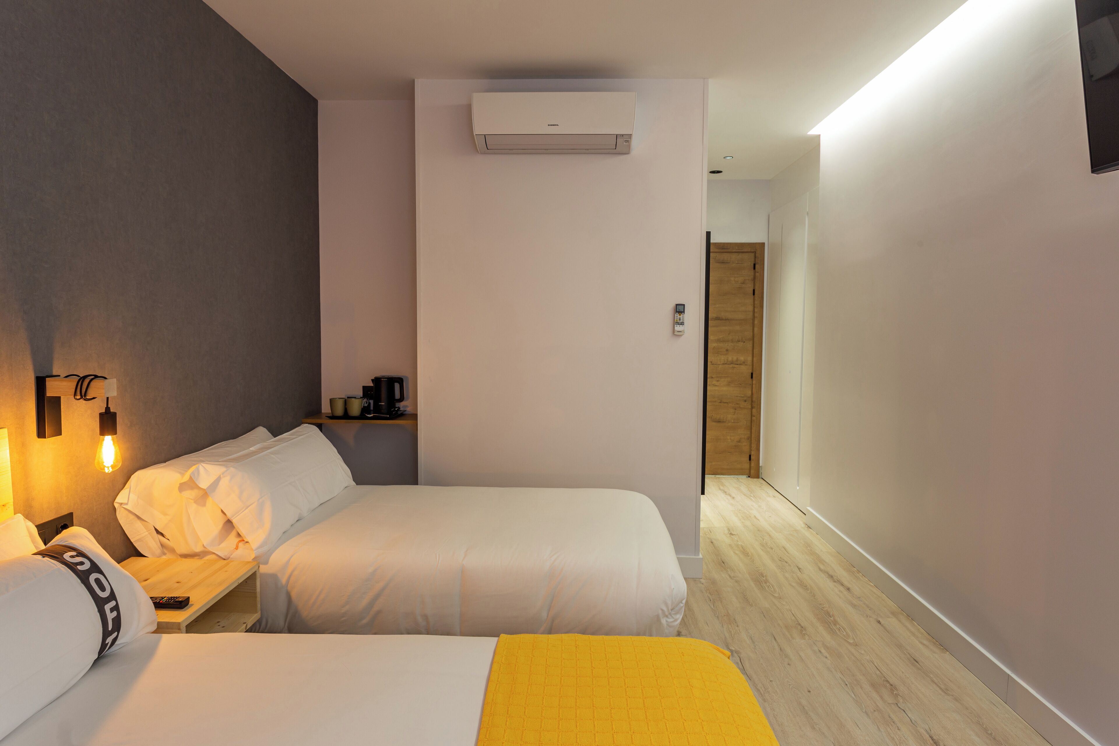 A hotel room with two beds, a bedside table, a lamp, an air conditioner, and a wooden door leading to another room.