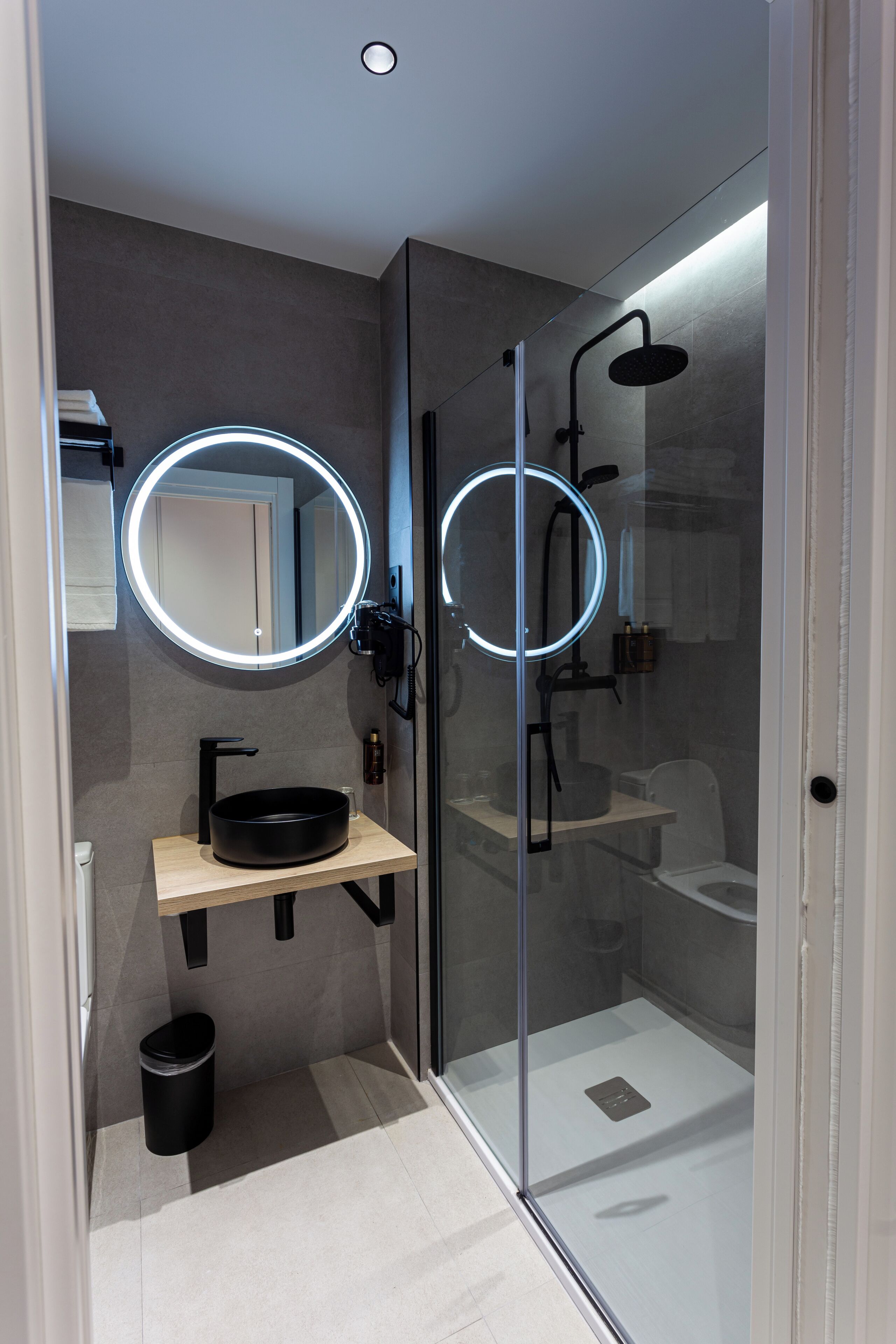 A modern bathroom with a glass-enclosed shower, a round mirror with underglow lighting, and a black sink.