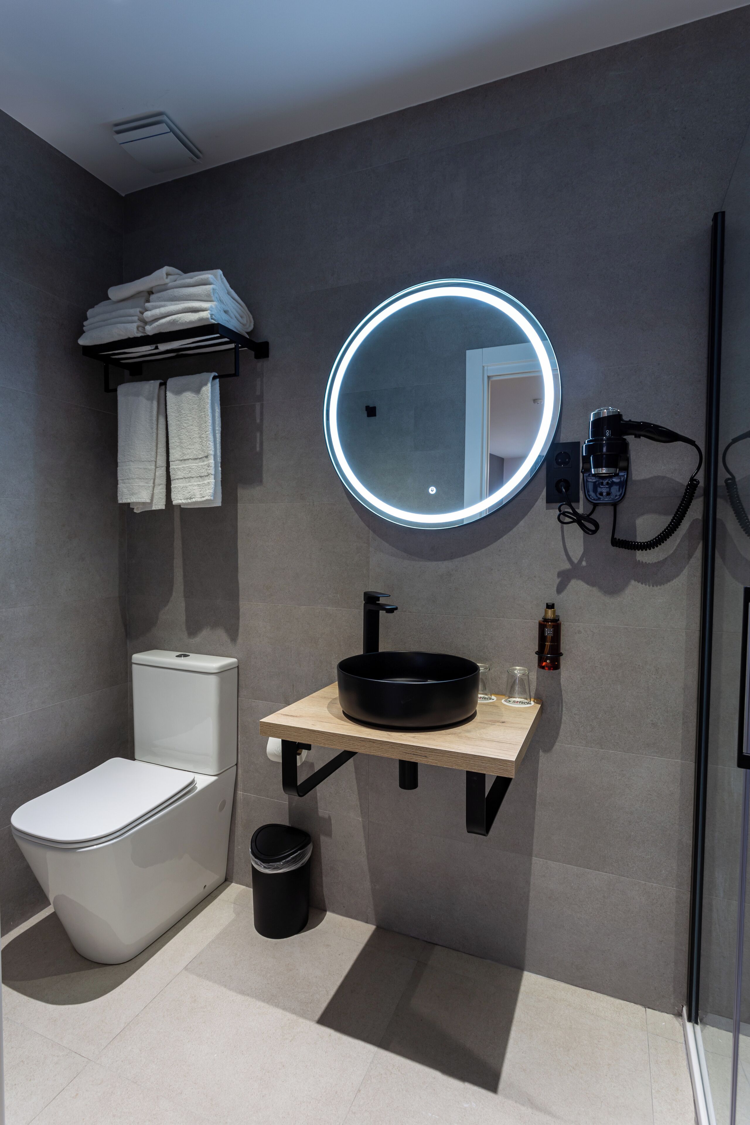A modern bathroom with a white toilet, a round mirror with a light, a black sink, and a towel rack.