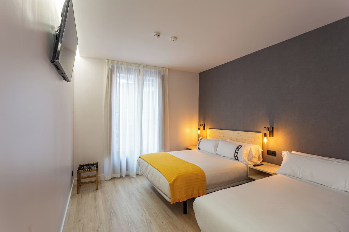 A hotel room with two beds, a TV mounted on the wall, a bedside table with a lamp, and a window with curtains.