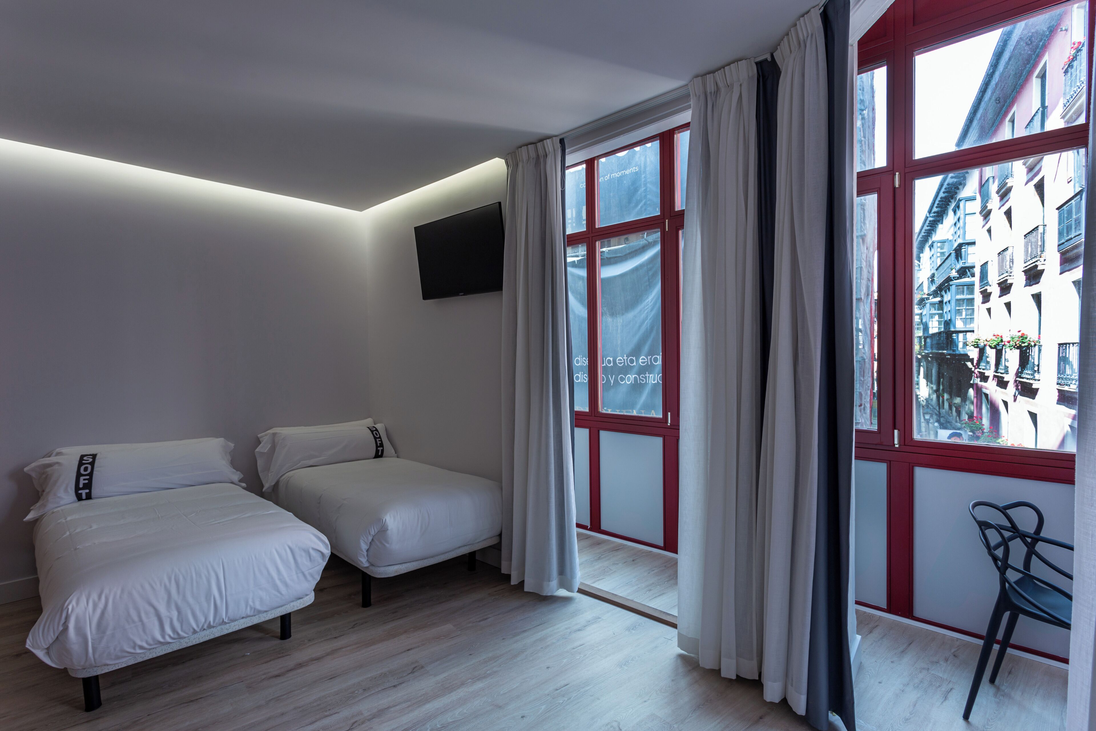 A bedroom with two beds, a TV mounted on the wall, and a large window with red frames.