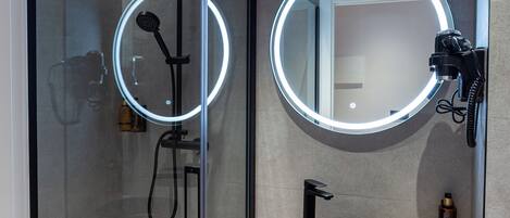 A modern bathroom with a glass-enclosed shower, a round mirror with LED lights, a black sink, and a wooden shelf.