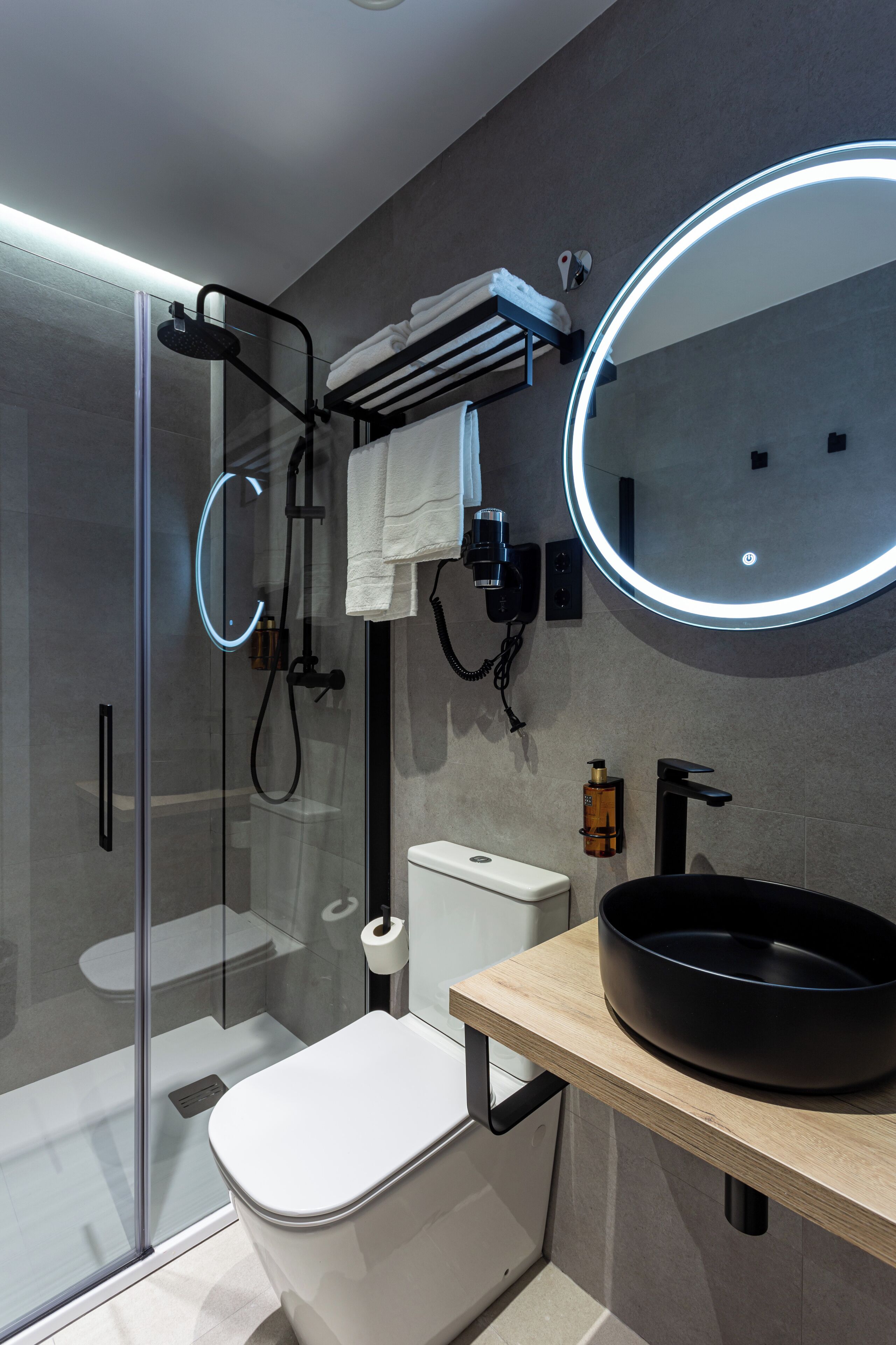 A modern bathroom with a glass shower enclosure, a toilet, a sink with a black basin, and a round mirror with a lighted frame.