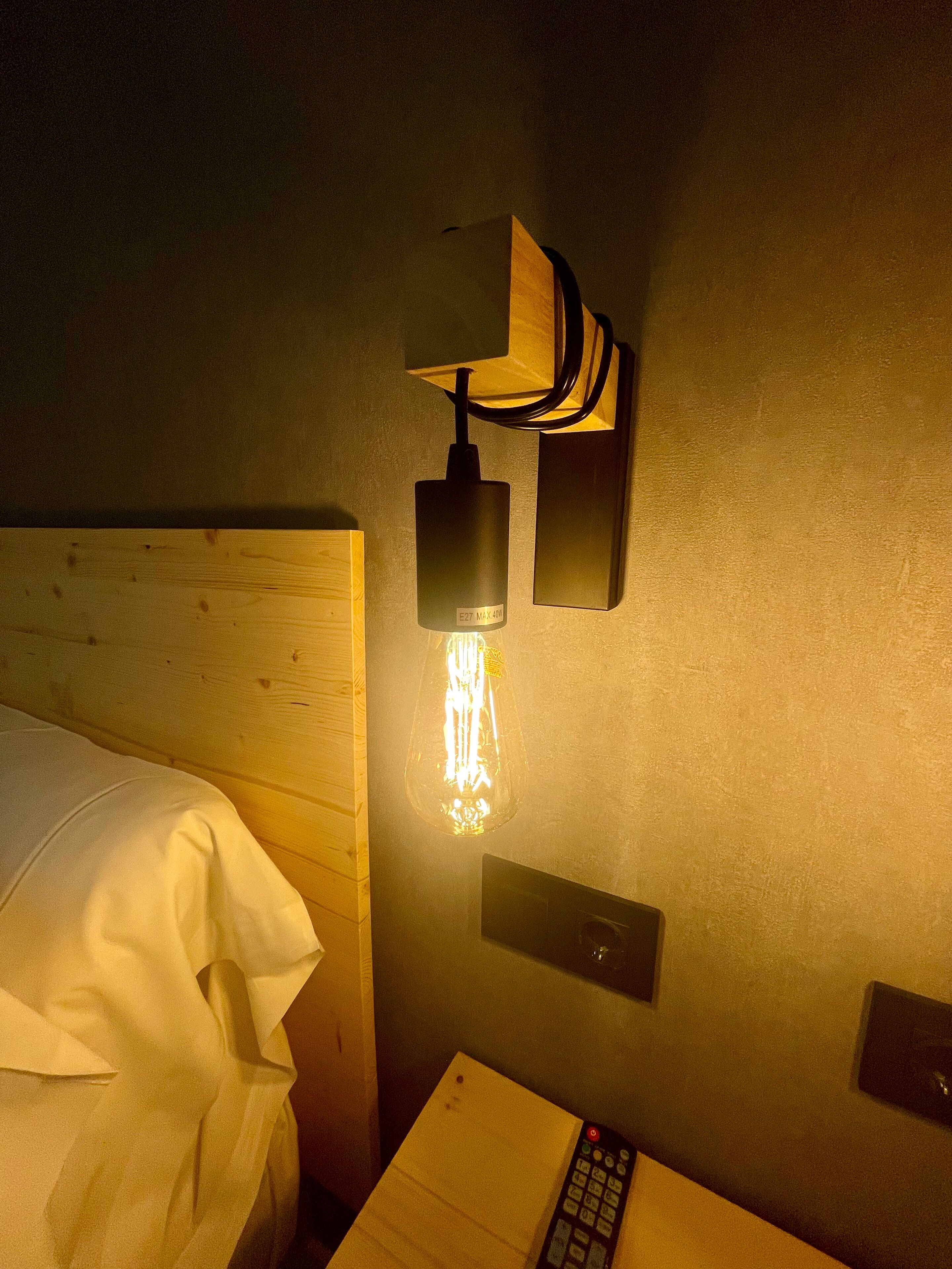 A hotel room with a wooden headboard, a bedside table with a remote control, and a wall-mounted light fixture with a clear glass bulb.