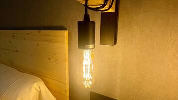 A hotel room with a wooden headboard, a bedside table with a remote control, and a wall-mounted light fixture with a clear glass bulb.