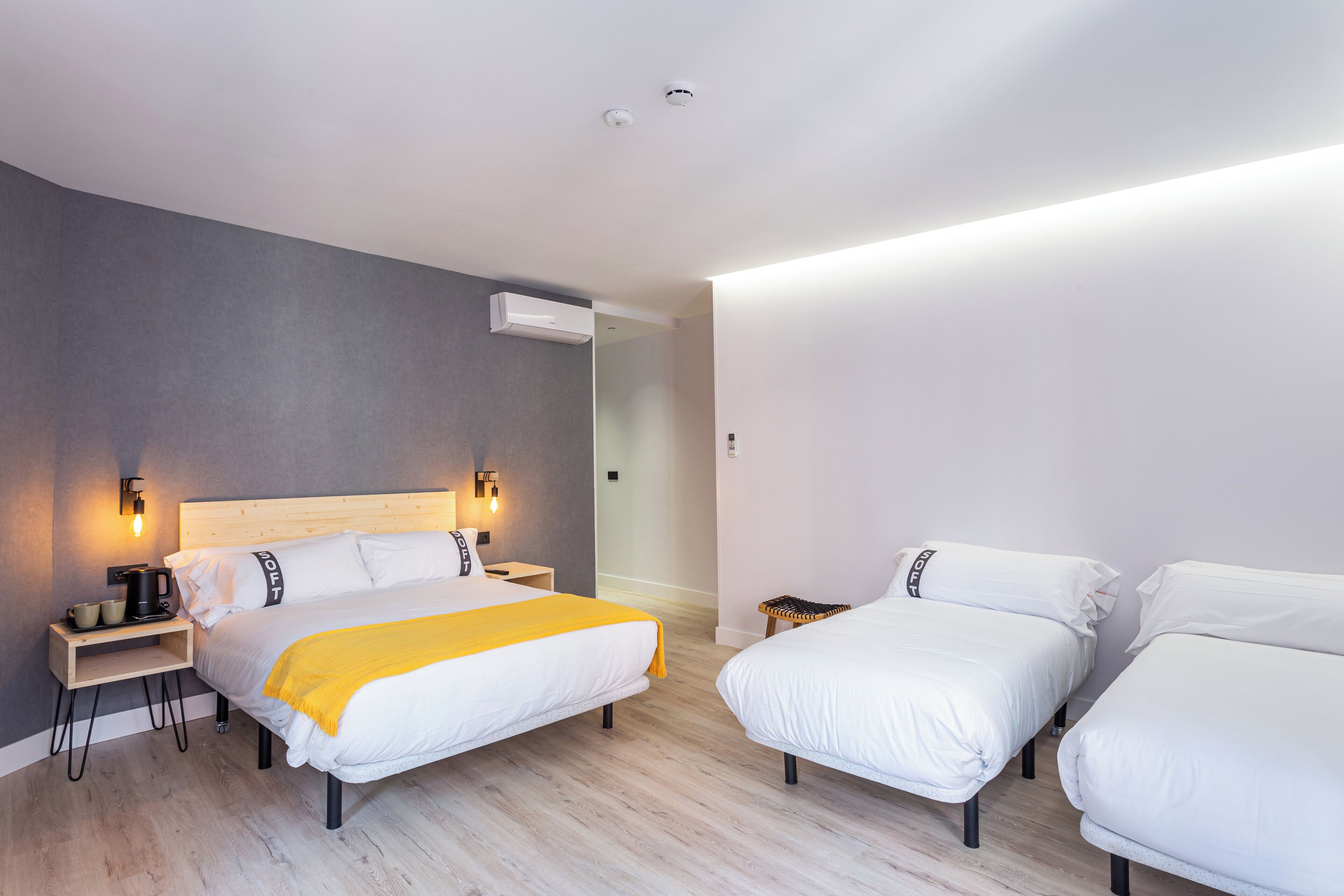 Moderni hotellihuone, jossa on suuri sänky, kaksi pienempää sänkyä, yöpöytä lampun kanssa ja seinälle asennettu ilmastointilaite.