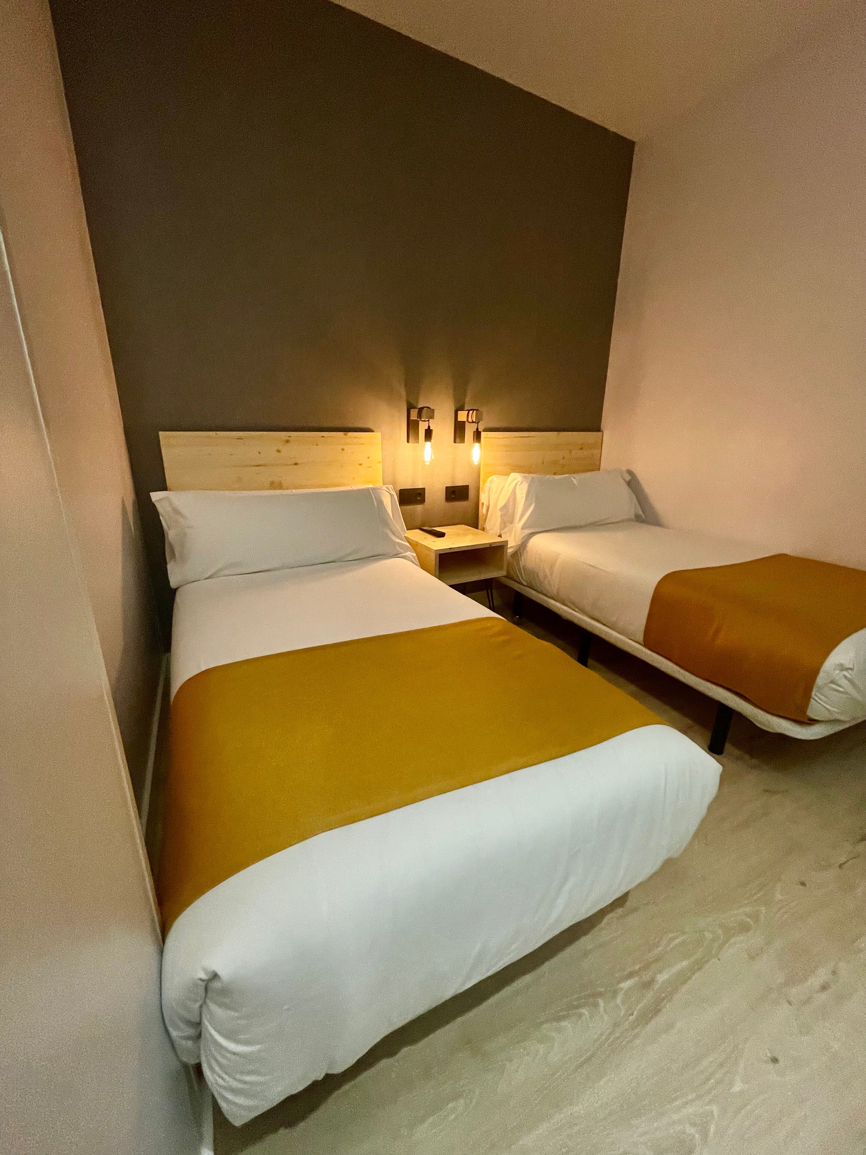 A hotel room with two beds, each with white bedding and a brown throw blanket. There are two wall-mounted nightstands with lamps.
