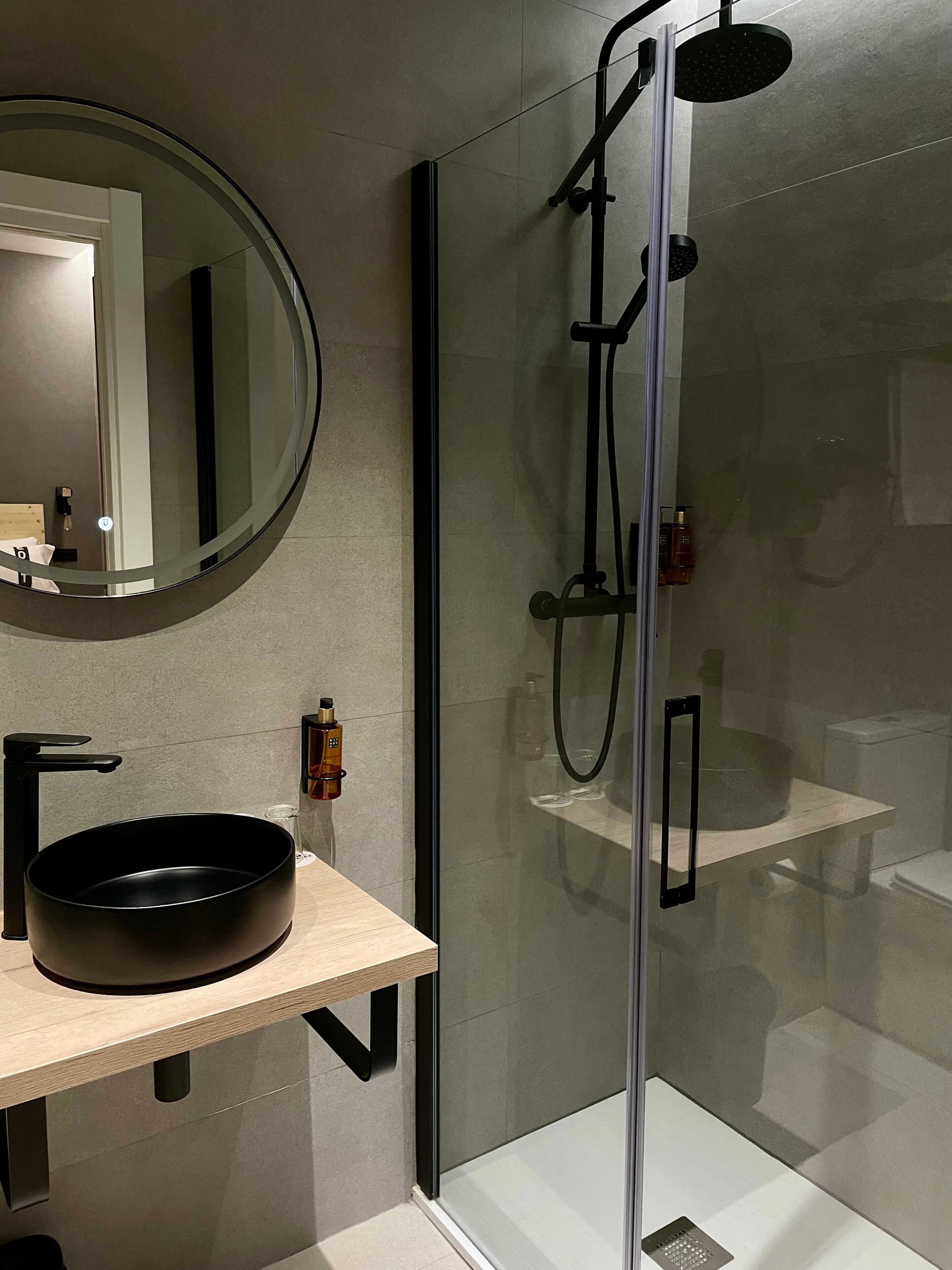 Un baño moderno con un espejo redondo, lavamanos negro y ducha con mampara de vidrio.