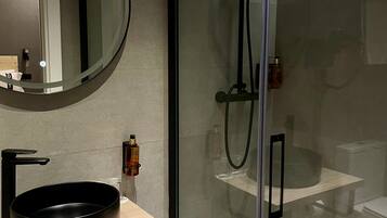 Un baño moderno con un espejo redondo, lavamanos negro y ducha con mampara de vidrio.