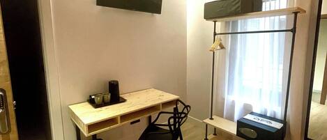 Una habitación pequeña y bien iluminada con un escritorio de madera, una silla negra, un televisor de pantalla plana montado, una ventana con cortina y un estante con una caja.
