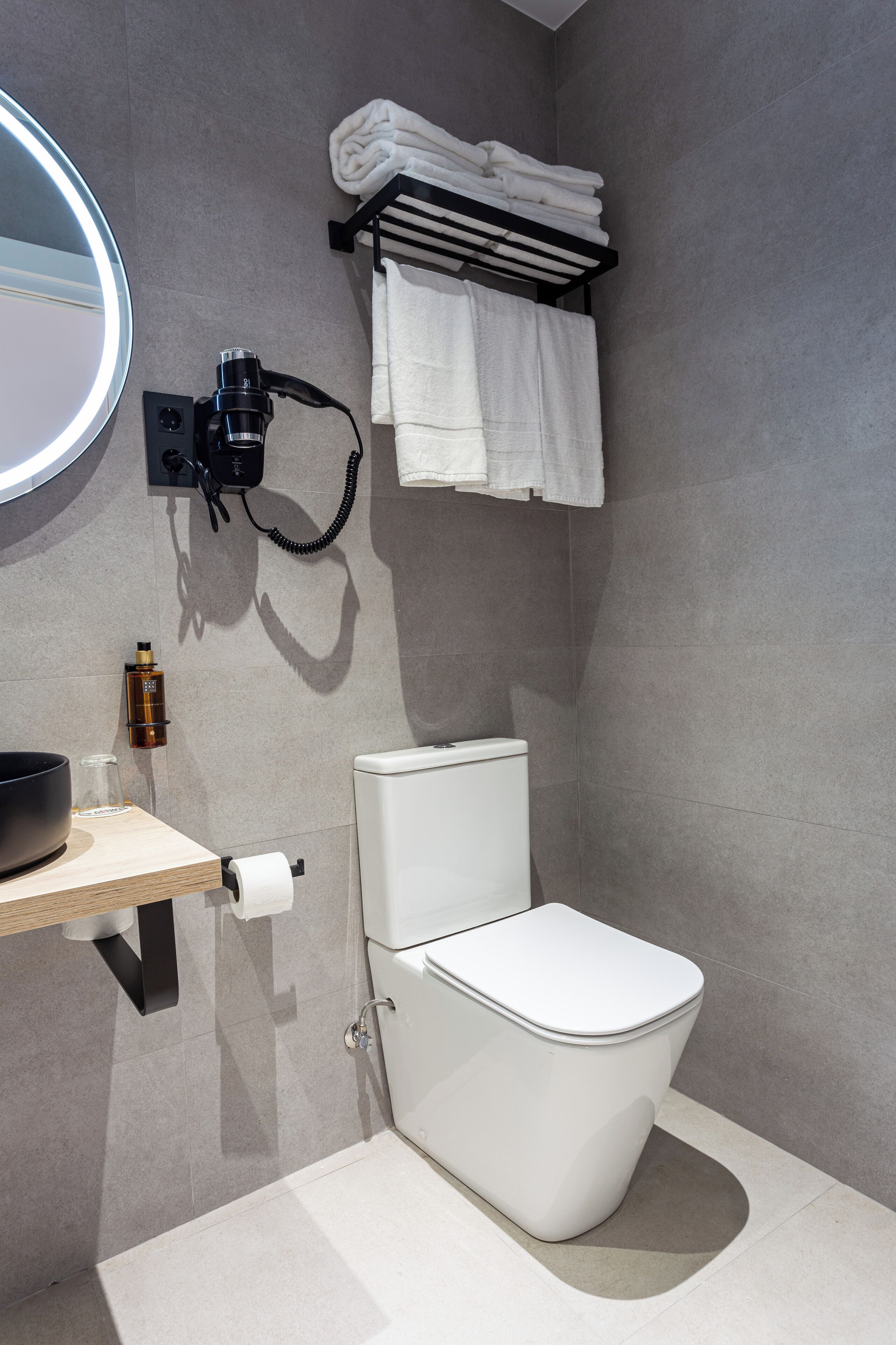 A modern bathroom with a toilet, a sink with a mirror, a towel rack with folded towels, and a bottle on the counter.