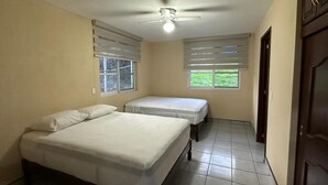 Quarto com duas camas, um ventilador de teto e janelas com cortinas.