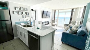 A modern kitchen with a built-in dishwasher, a large island with a sink, and a dining area with a blue sofa. - Gone Coastal/ Prime Location (Myrtle Beach)