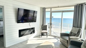 A modern living room with a flat-screen TV mounted on the wall, a fireplace, comfortable seating, and a large sliding glass door leading to a balcony with a view of the sea. - Gone Coastal/ Prime Location (Myrtle Beach)