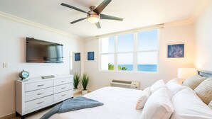 Un dormitorio con cama, ventilador de techo, televisor, cómoda, ventana con vistas y una obra de arte colgada en la pared.