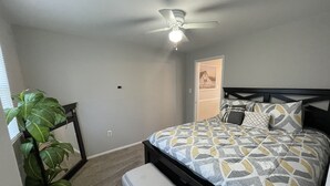 A bedroom with a bed, a ceiling fan, a potted plant, and a door leading to another room.