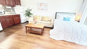 Habitación de hotel compacta con kitchenette, sofá con cojines, cama con edredón blanco y una pequeña mesa de madera con una planta.