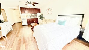 A bedroom with a bed, a nightstand, a dresser, a vanity, a ceiling fan, a kitchen area with cabinets, and a dining table.