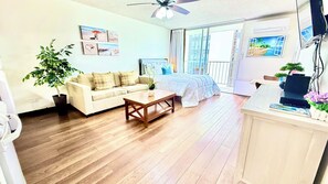 A living room with a sofa, a coffee table, and a bed with a blue bedspread. There are wall art pieces and a ceiling fan.