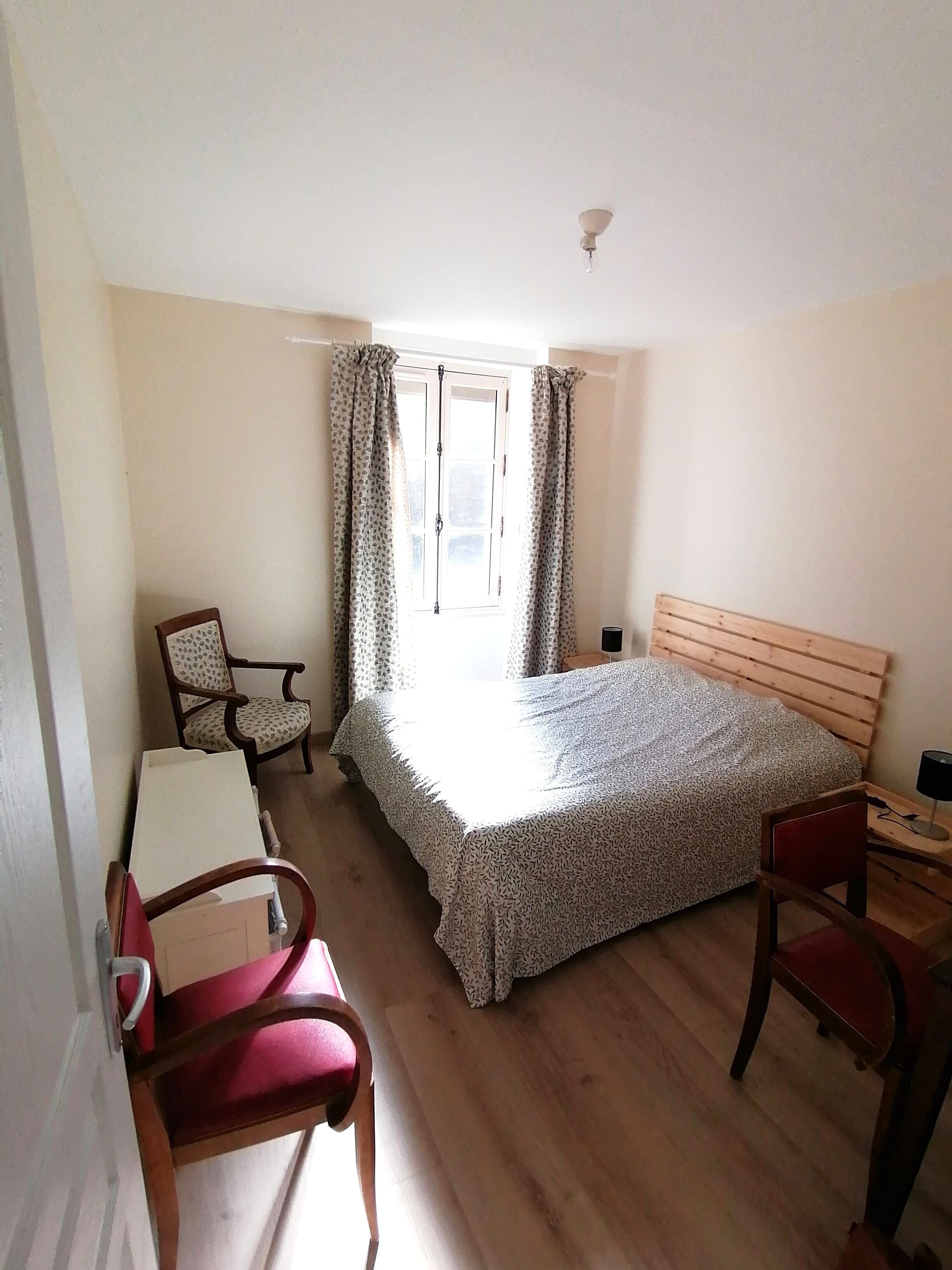 A small, well-lit bedroom with a single bed, two chairs, a desk, and a window with curtains.