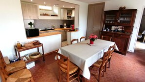 Un coin repas avec une table dressée pour quatre personnes, entourée de chaises en bois. On aperçoit, au fond, un espace cuisine avec des placards et un plan de travail.