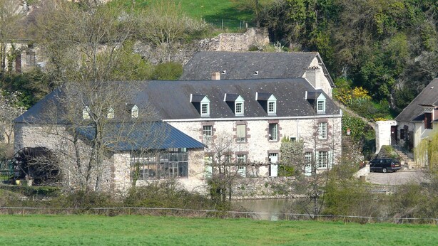 Un grand bâtiment en pierre, doté de nombreuses fenêtres et d’un toit en pente, entouré de verdure et bordé d’un petit plan d’eau.