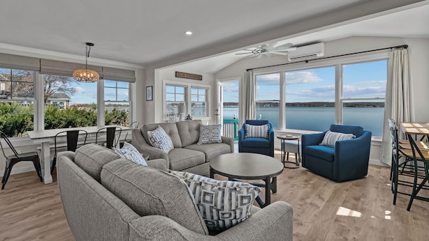 A spacious living room with a large window overlooking a body of water, a ceiling fan, and a dining area with a table and chairs.