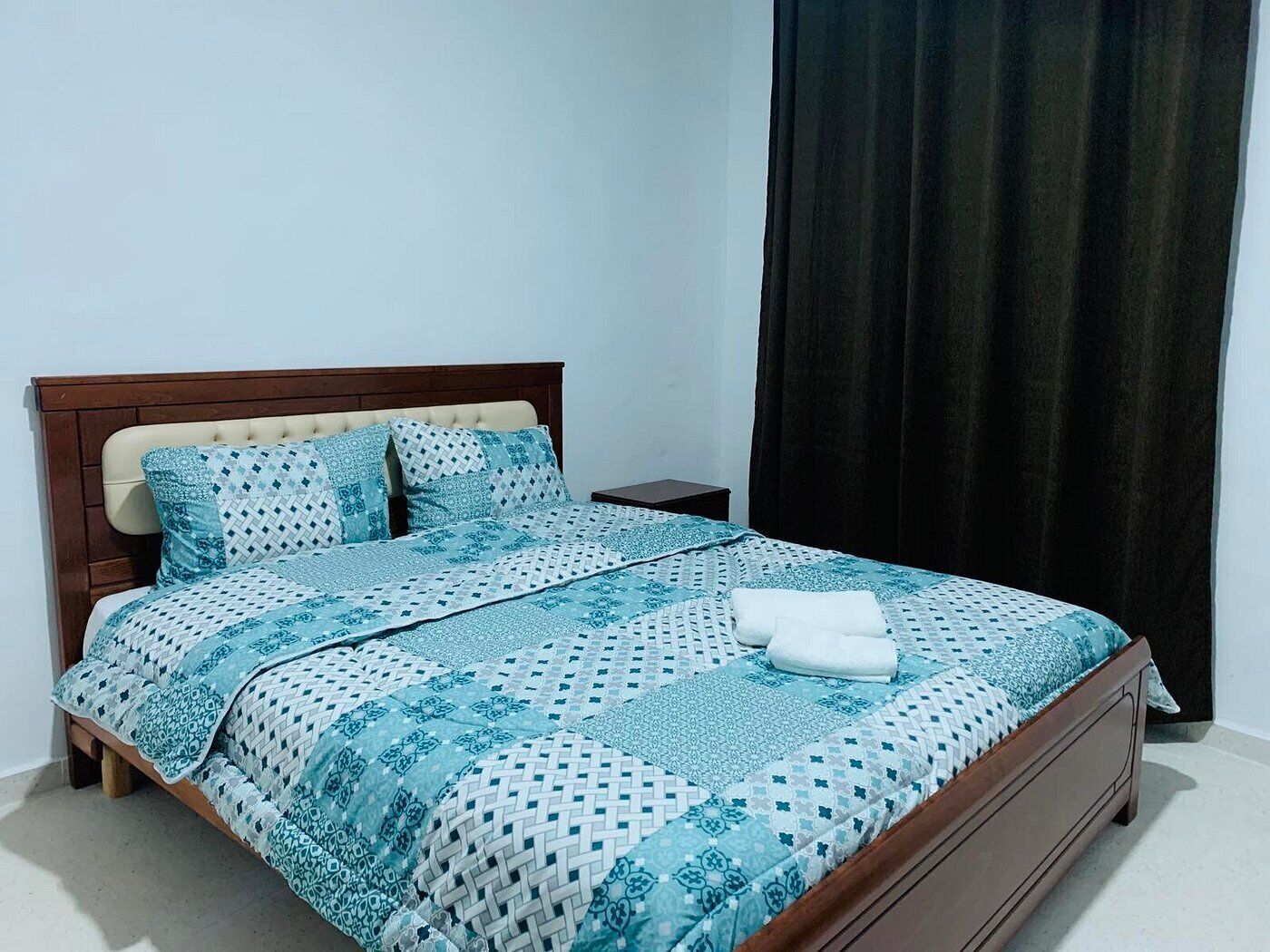 A neatly made bed with a patterned bedspread and pillows, a wooden headboard, a small bedside table, and a dark curtain.