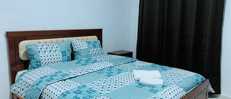 A neatly made bed with a patterned bedspread and pillows, a wooden headboard, a small bedside table, and a dark curtain.