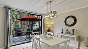 A dining area with a white table, chairs, a red umbrella, and a large window with a view of trees and a balcony. - Updated Beach Villa  2 bd 2 ba  just steps 2 sand! (Hilton Head Island)