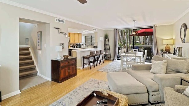 A spacious living room with a large sofa, a coffee table, and a dining area with a table and chairs. There is a kitchen area with a bar and stools, and a staircase leading to another level. - Updated Beach Villa  2 bd 2 ba  just steps 2 sand! (Hilton Head Island)