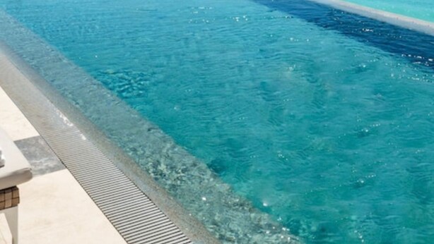 A long swimming pool with clear blue water, adjacent to a modern building with large windows overlooking the ocean.