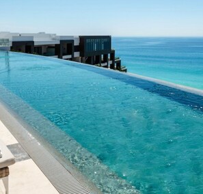 En lang swimmingpool med klart, blåt vand, ved siden af en moderne bygning med store vinduer med udsigt over havet.
