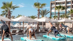 A poolside area with people doing yoga on floating platforms, palm trees, and a hotel building in the background.
