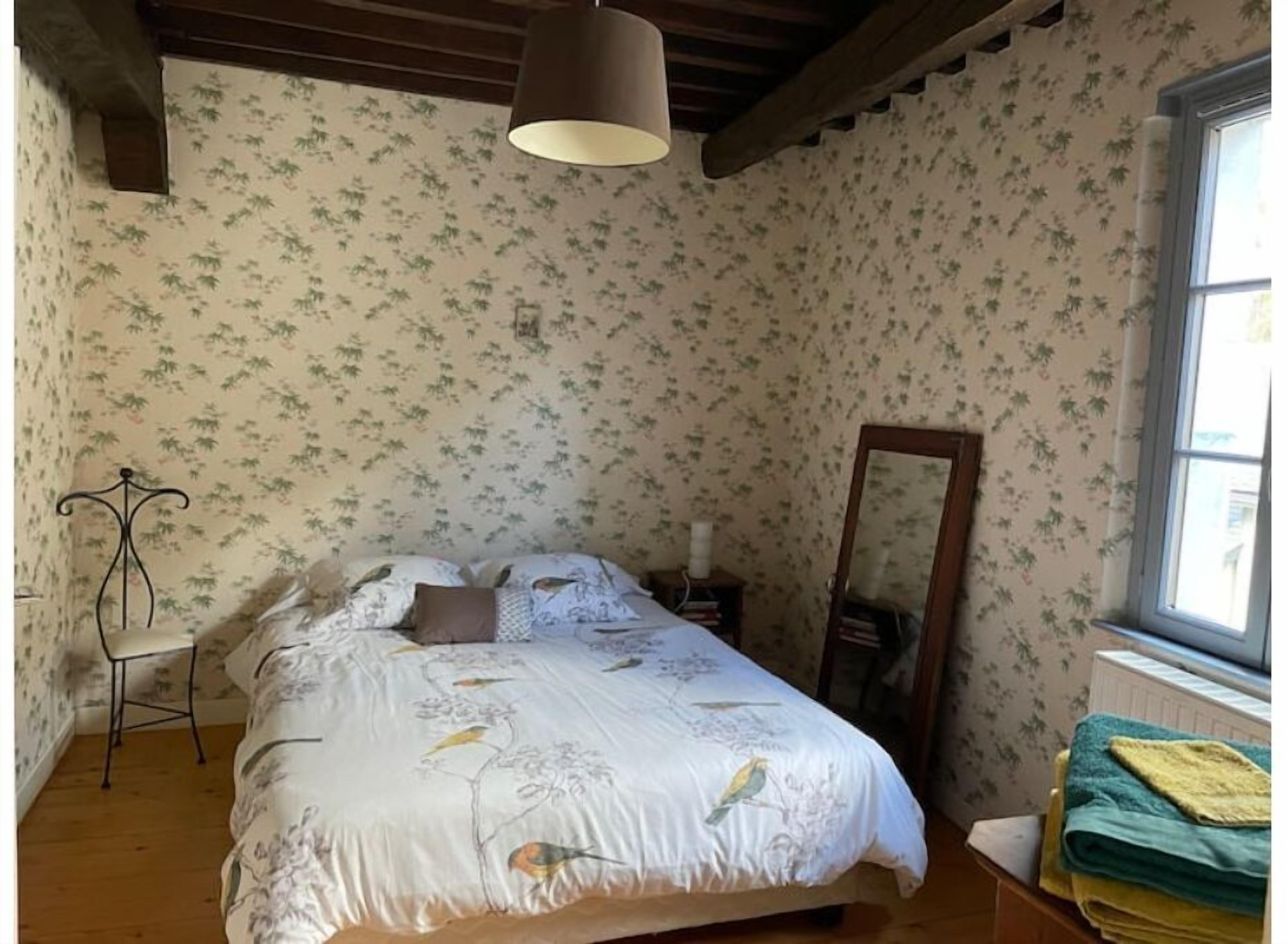 Une chambre avec un papier peint à motifs floraux, un lit recouvert d’une courtepointe blanche ornée d’oiseaux, une table de chevet avec une lampe et une fenêtre donnant sur un beau paysage.