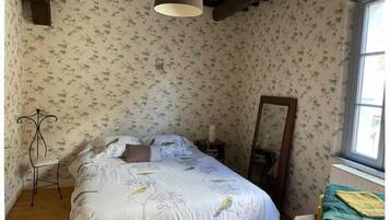 Une chambre avec un papier peint à motifs floraux, un lit recouvert d’une courtepointe blanche ornée d’oiseaux, une table de chevet avec une lampe et une fenêtre donnant sur un beau paysage.