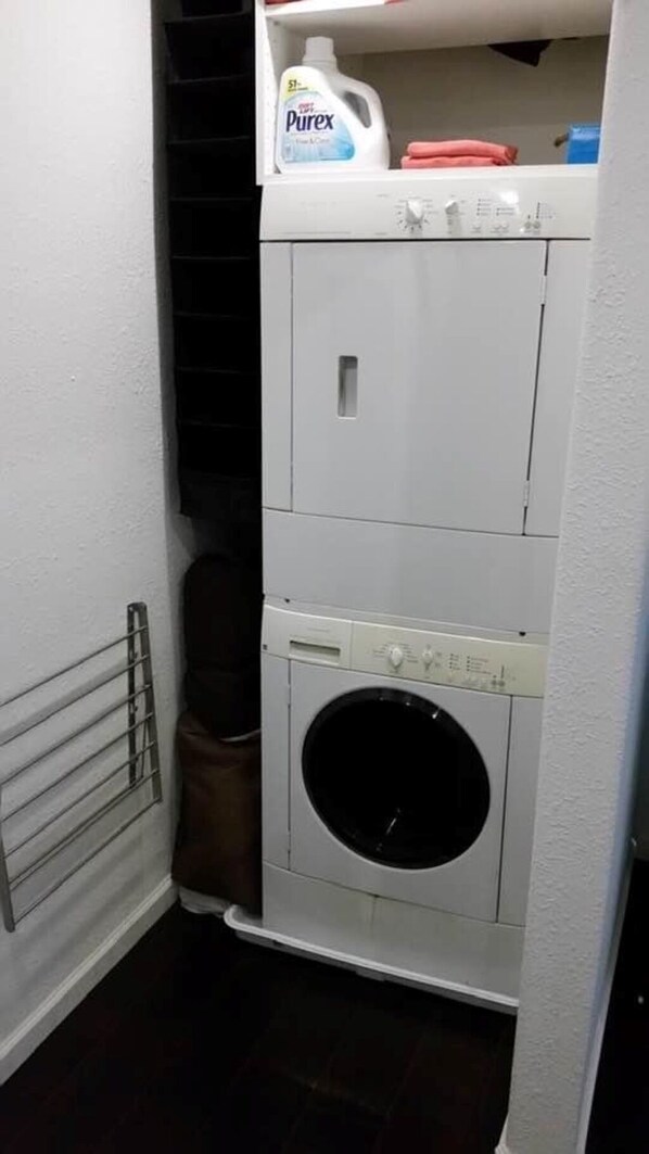 A compact laundry area with a washing machine and a dryer, a bottle of Purex detergent, and a brown storage bin.