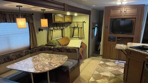 Interior compacto de una caravana con comedor, asientos, kitchenette y televisor.