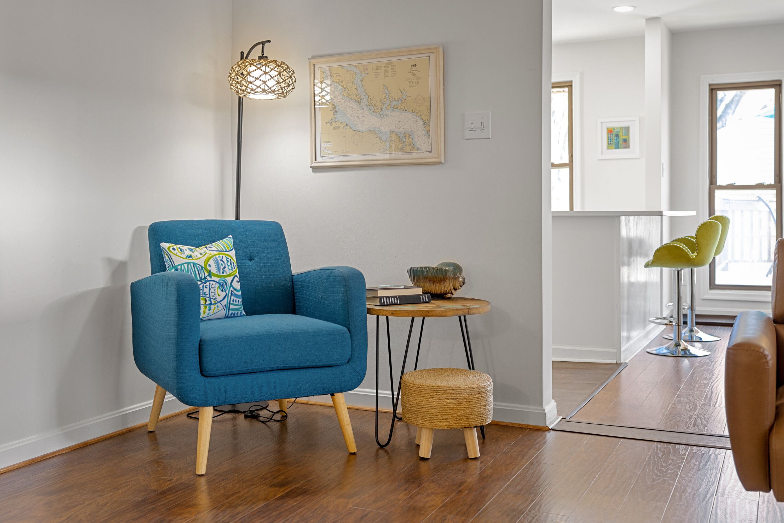 Una sala de estar moderna con un sillón azul, una mesita redonda con un libro y un cuenco decorativo, un taburete tejido y una lámpara de pie.