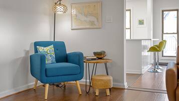 Una sala de estar moderna con un sillón azul, una mesita redonda con un libro y un cuenco decorativo, un taburete tejido y una lámpara de pie.