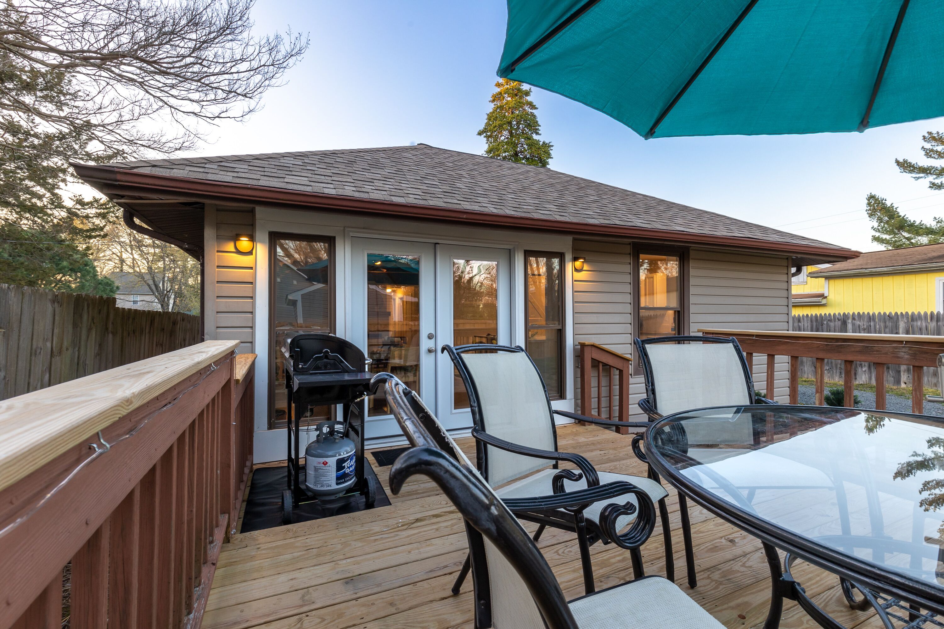 A deck with a glass table and chairs, a barbecue grill, and a wooden fence.