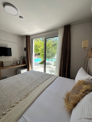 Habitación de hotel con una cama grande, televisión, escritorio con hervidor y vista a una piscina por una puerta de cristal.