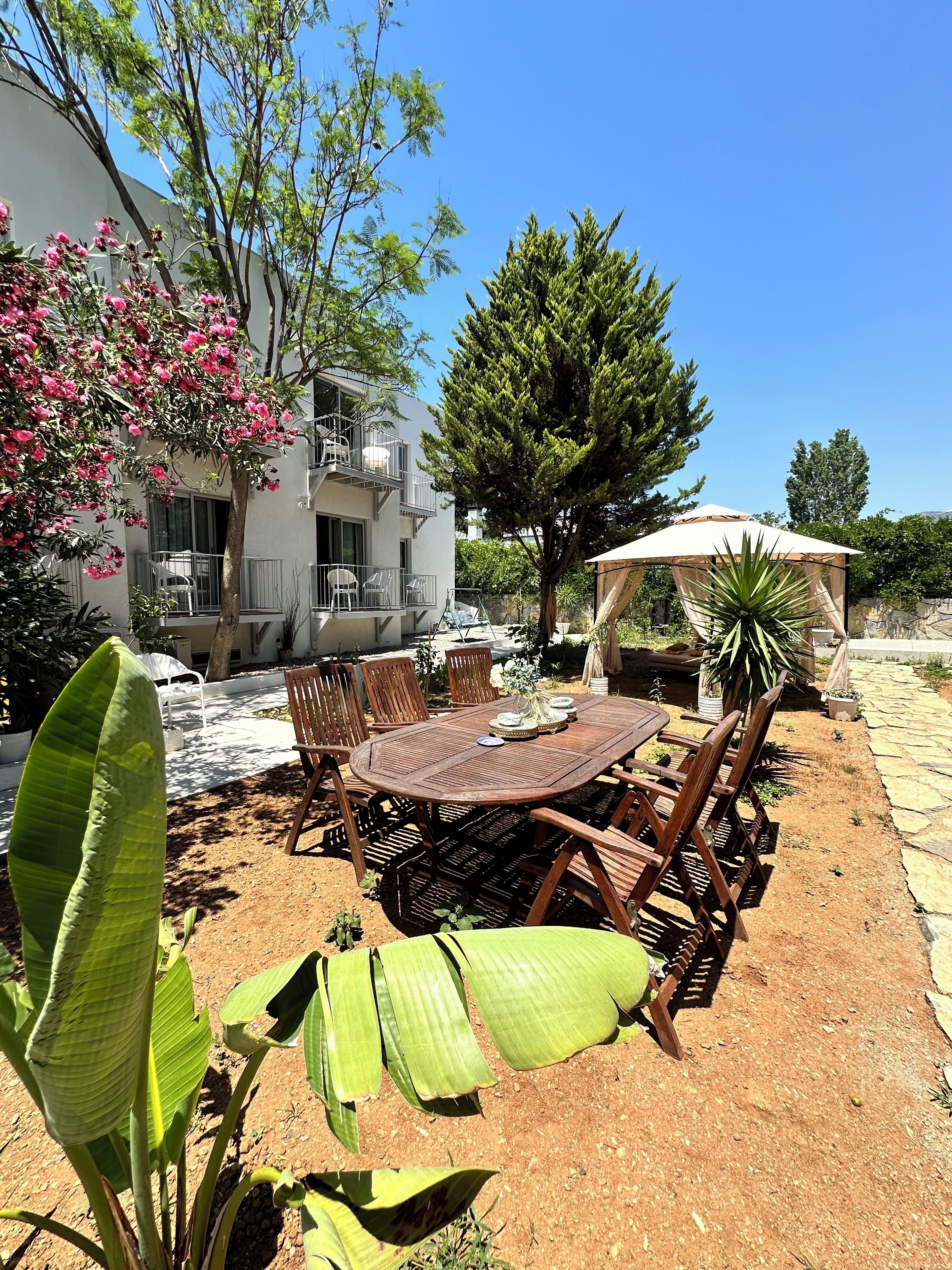 An outdoor patio with a round wooden table and chairs, surrounded by plants and a large umbrella.